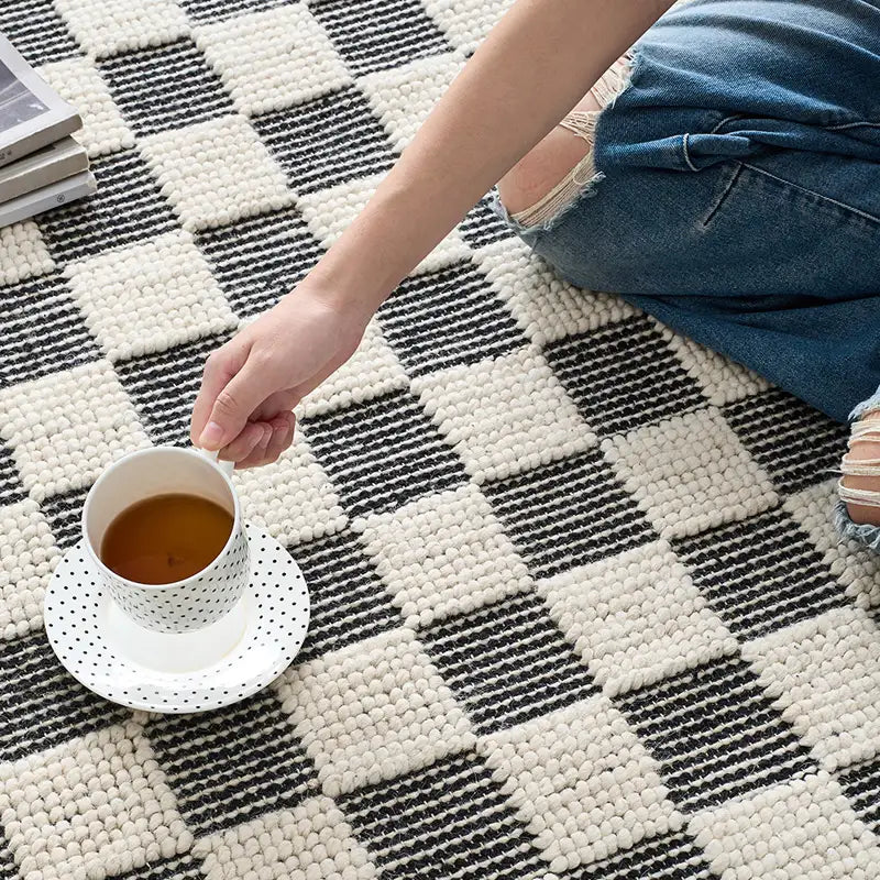 Close-up of a black and white checkered wool rug, featuring a hand holding a cup of tea, showcasing its soft texture and cozy design #color_Black & White Check