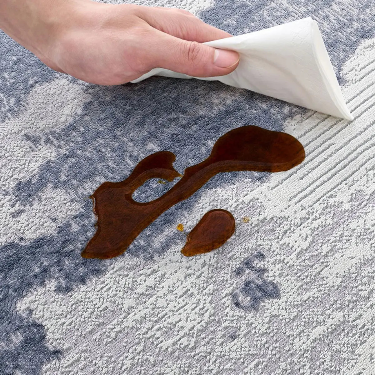 Blue-gray abstract area rug spill-resistant detail with a paper towel wiping an everyday liquid spill from the textured surface.