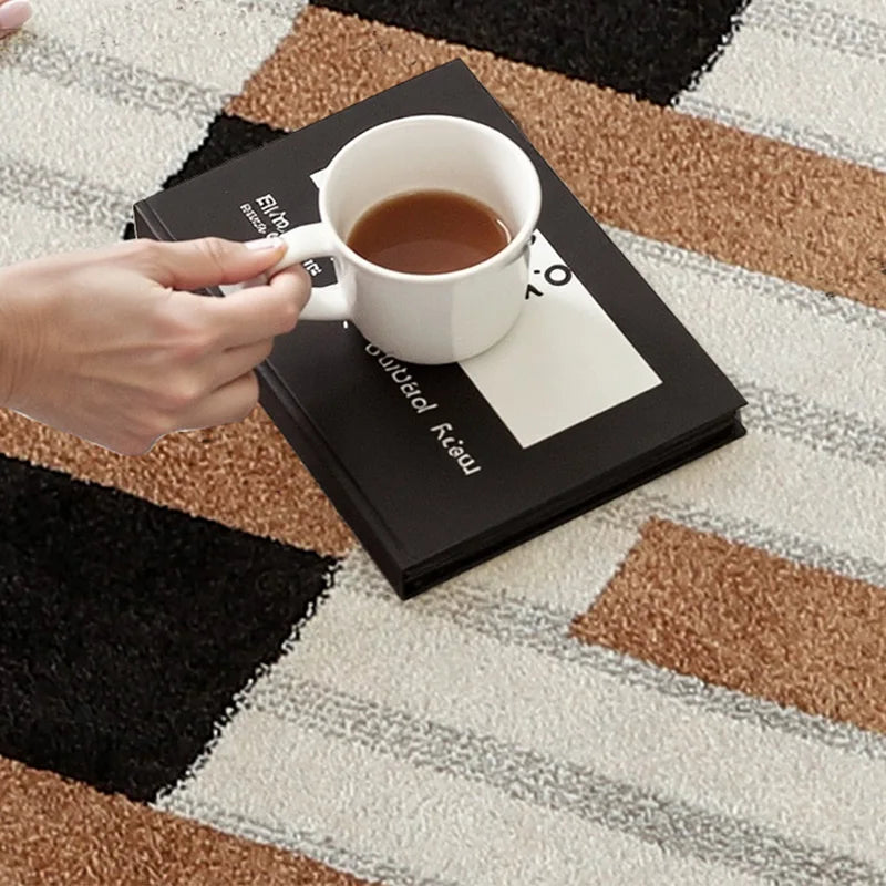 Hand holding a coffee cup resting on a book placed on a geometric checkered rug in warm brown and black tones